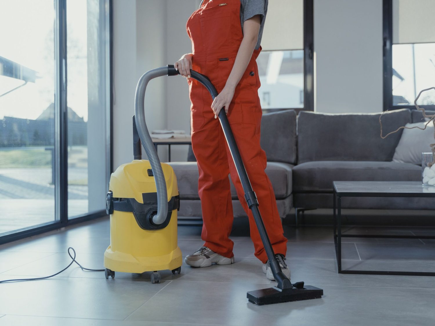 A professional cleaner in red coveralls vacuuming a modern living room with a yellow vacuum cleaner.
